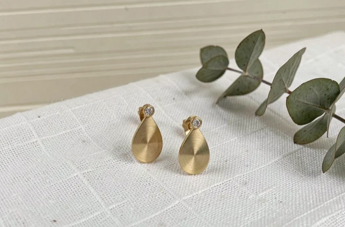 Boucles d'oreilles Torrente en or brossé avec accent diamant présentées sur un fond lin blanc dans un style éditorial épuré
